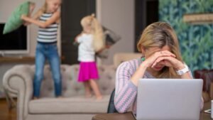 Mujer estresada frente al ordenador con la cabeza entre las manos, mientras dos niños juegan detrás en un ambiente caótico. La imagen representa cómo manejar el estrés y transformar la tensión en claridad emocional en situaciones cotidianas.